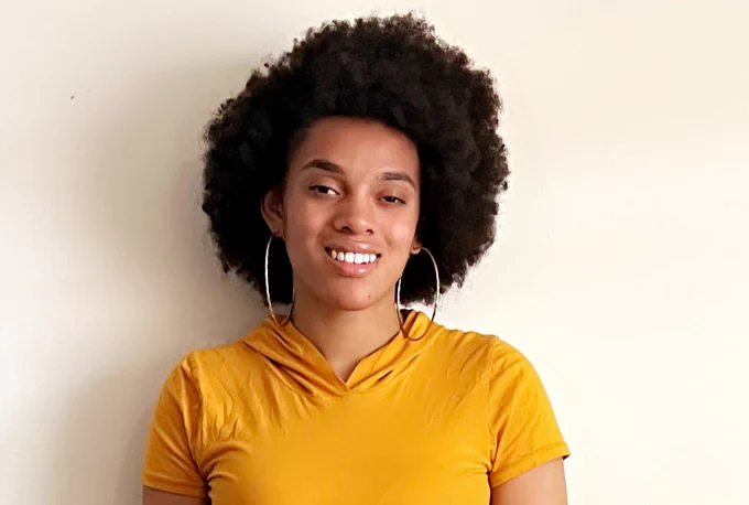 This photo shows Taty standing in front of a white wall. She is wearing a yellow shirt and silver hoop earrings, and she is smiling at the camera.
