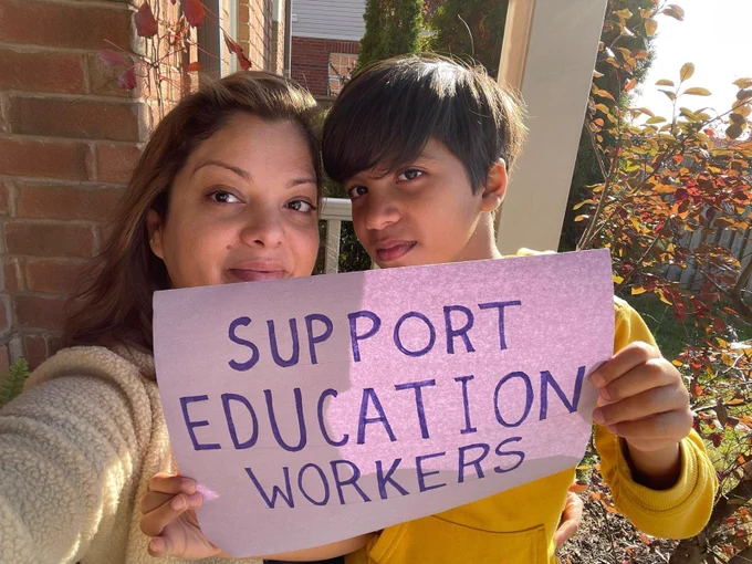 Woman and boy hold handmade sign supporting education workers. Standing in front of home with fall foliage in the background.
