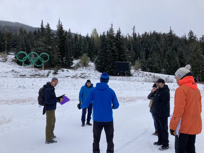 A group of cold-looking people standing in a circle in the snow. In the background is some coniferous forest and a big green sculpture of the Olympic Rings.
