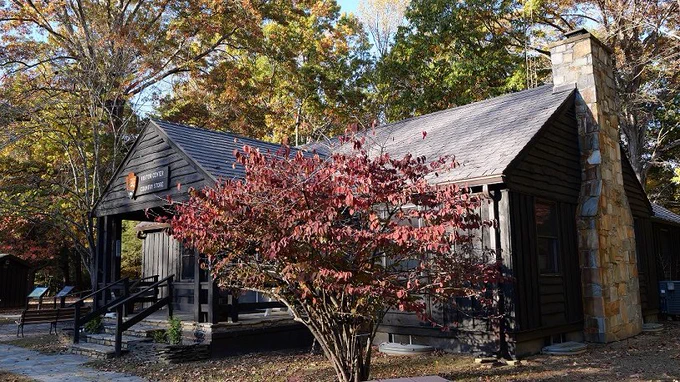 Brown wooden cabin visitor center with stone chimney and porch in a fall forest with a large red leafed bush in front
