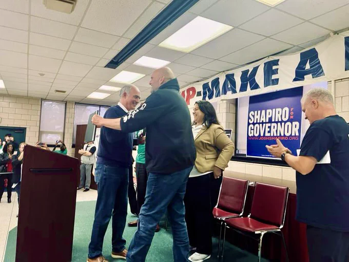 Senator Casey and John Fetterman start to hug as Casey, who just finished speaking, passes his mic to Fetterman.