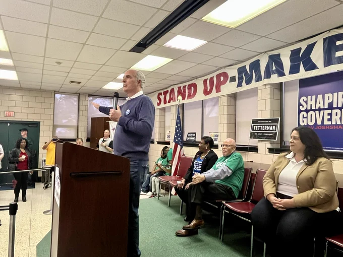 Senator Casey, raising one hand, speaks into a mic at a podium in front of Fetterman and Shapiro signs.