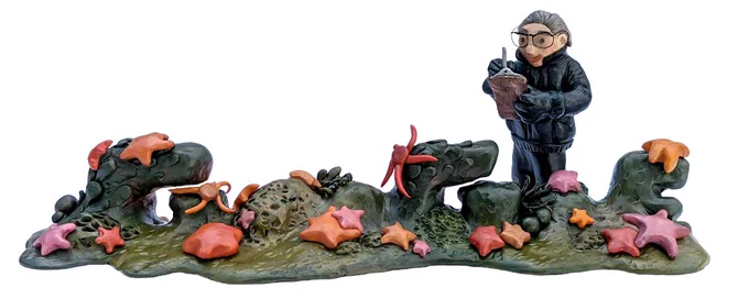 Clay sculpture of Irene Bernasconi standing on the sea floor which is abstractly formed into the "GOOGLE" letters with starfish scattered thorughout. Irene has short grey hair, is wearing an all black outfit, has glasses, and is holding a clipboard. 