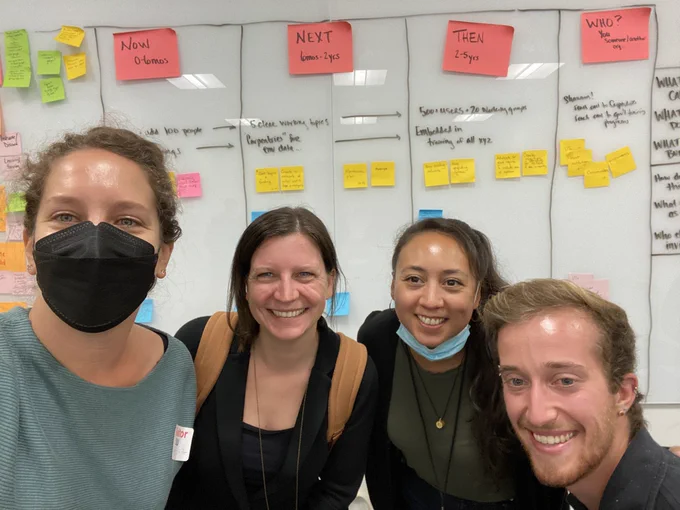 Four people stand in front of a white board with writing on it.