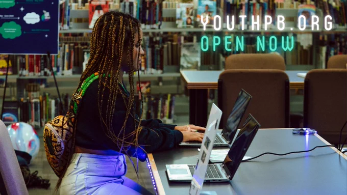 Glowing white and teal text “YouthPB.org Open Now” and a young person with long brown braids and a backpack sitting at a desk in the library using a laptop computer.