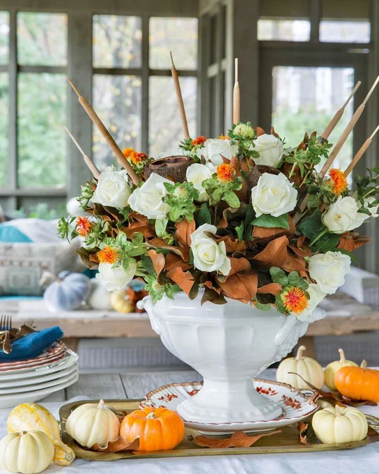 In a large white vase, a centerpiece of white roses, orange safflowers, cattails, and dried magnolia leaves is surrounded by mini pumpkins.