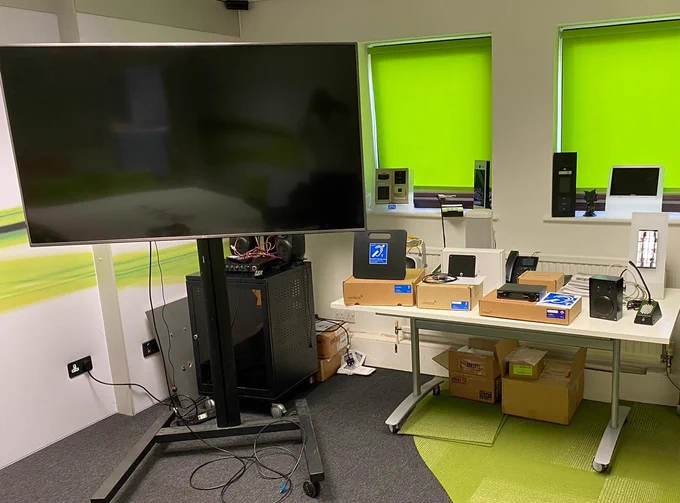 A large TV screen in a training room. A range of Contacta products are set out on a table next to it, infront of lime green blinds.