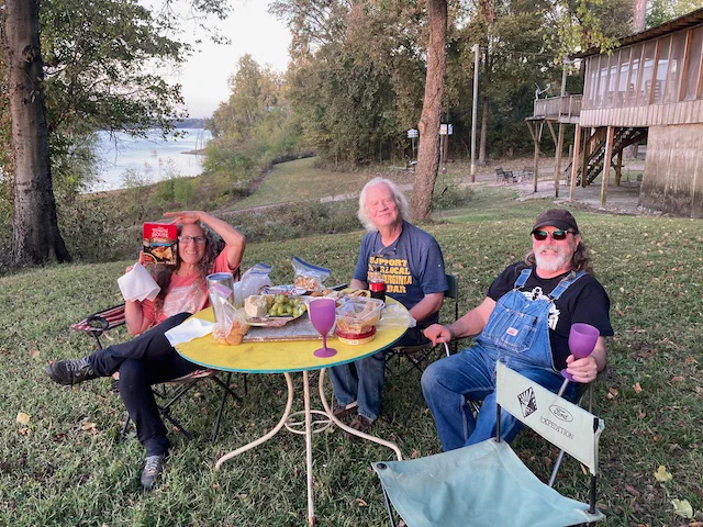 Carol Vincent, Gary Vincent, and Adrian Kosky, seated at an outdoor table full of snacks and beverages at sunset on the banks of Desoto Lake, Rena Lara, Mississippi, USA.
