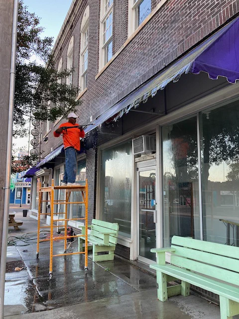 Ben Williams standing on scaffold, pressure washing purple awnings above windows and green benches at The Holy Moly building in Clarksdale, Mississippi, USA. Old masonic temple two-story building.