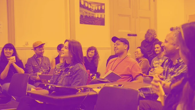 Common Field Network members attend the 2019 Convening in Philadelphia, PA. Participants sit in a classroom, oriented towards the front. They review literature and engage with the discussion, joyfully, smiling. In the middle, a young person in a red shirt and black baseball hat looks forward mirthfully. In front of him, a young person with long brown hair wears a denim jacket and does the same. They sit beside a table around which many other participants are gathered and upon which a conference-style telephone rests.. Behind them, a wall with a large wooden double door. Convening participants sit in chairs along the wall. Above them hangs a photograph of the Friends Center meeting space, a large wooden Quaker meeting room. The image has a gold and purple graphic wash overlaid on the photograph.