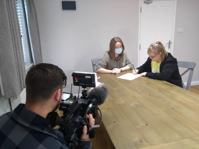 Two people sit at a table writing on a piece of paper while being filmed by a camera operator