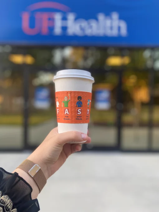A coffee cup is held up in front of the entryway to a building. The banner above the door reads UF Health. The cup has a sleeve with the steps of what to do to evaluate a stroke and what to do next, FAST. If you see or experience face drooping, arm weakness, or speech difficulty, it's time to call 911.