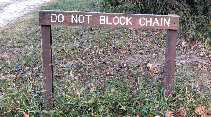 Wooden sign in field next to a gravel road. The sign reads, “DO NOT BLOCK CHAIN.”