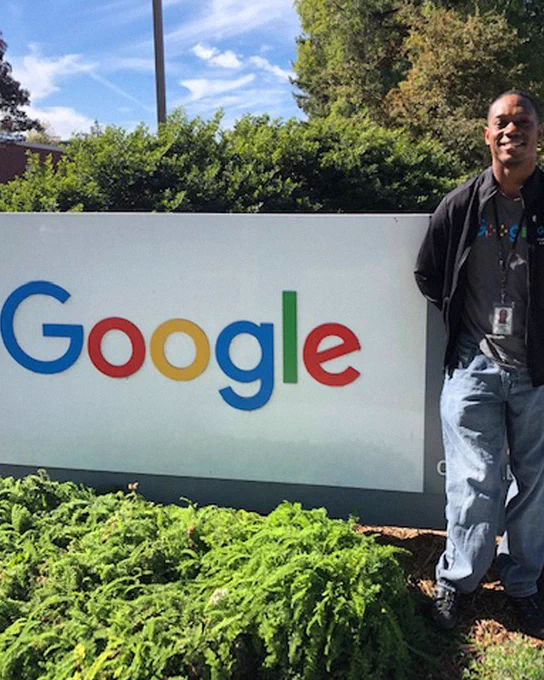 Jerry smiling and leaning against a Google building sign, with his arms folded, wearing jeans and a dark jacket.