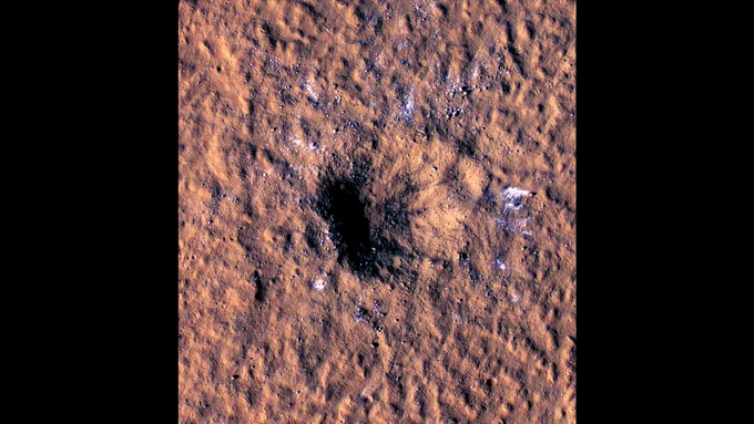 A rust-colored image shows a black hole that is an impact crater on the surface of Mars as seen from an orbiter in space. The black hole is deep and slightly oblong shaped. The texture of the surface of Mars looks pockmarked and there are light-blue splotches around the crater. Those splotches are ice that was splattered around the crater following the meteroid crash.