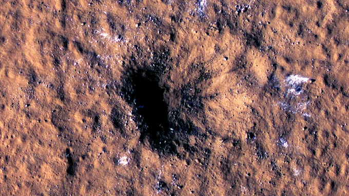 A close-up picture of the reddish-brown surface of Mars with a large crater captured by the Mars Reconnaissance Orbiter.  The crater is in the middle of the image and has darker material, almost black inside due to the impact which excavated material from within, exposing chunks of water-ice shown as bluish-white around the edges of the crater.