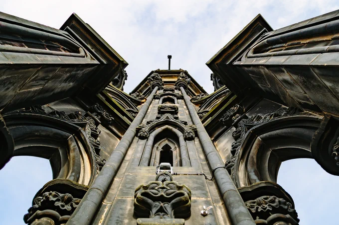 A view looking directly up at the Scott Monument in Edinburgh. The view has two arches that are part of flying buttresses on the left and the right. The center is a tower that continues on. The tower is made of stone and is filled with Gothic carvings and adornments: faces, flowers, plants, depictions of animals, etc.