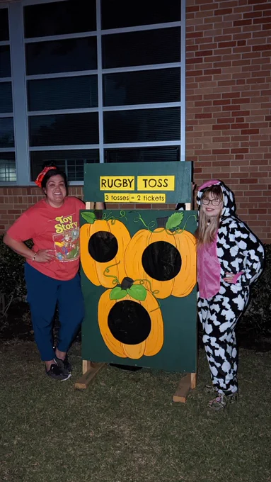 Ms. Erica and Ms. V standing on either side of a rugby toss game, painted with pumpkins, outside during Fall Festival.