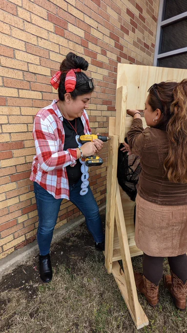 Ms. Erica and Ms. Mirna outside. Ms. Erica is using a drill to attach a sign to a wooden board. Ms. Mirna is holding the sign up.