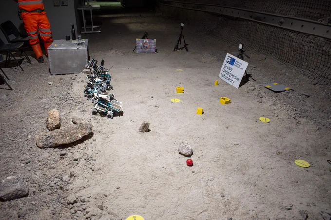 An image of Remote3 robots in Boulby Underground Laboratory. Four rovers can be seen with obstacles and things to go retrieve on the floor of the mine. The legs of a person in orange high-vis can be seen in the top left corner 