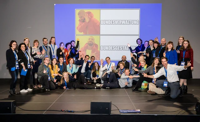 Eine große Gruppe Menschen auf der Bühne das ganze Team Tech4germany 2022. Hinter ihnen an der Wand ein Meme. Bundesverwaltung = Nein, Bundesgestaltung = Fein.