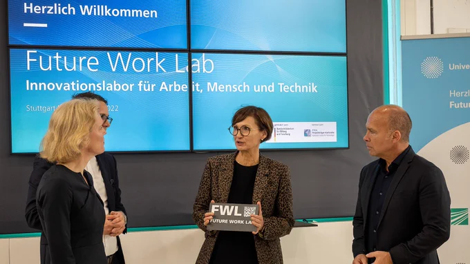 Forschungsministerin Bettina Stark-Watzinger im Gespräch mit Mitarbeitern des Future Work Labs. Sie hält eine Plakette mit der Aufschrift „Future Work Lab“.
