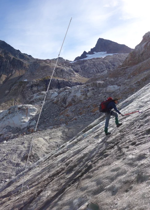 Researcher on slope of glacier with tall stake behind him and exposed mountain in background
