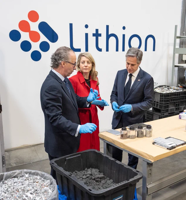 Secretary Blinken and Foreign Minister Melanie Joly listen as a gentleman shows them recyclable materials in bins and containers on a table at Lithion in Montreal. A white wall that reads "Lithion" sits behind them.