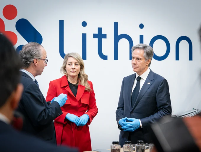 Secretary Blinken and Foreign Minister Melanie Joly watch as a gentleman shows them recyclable materials in bins and containers on a table at Lithion in Montreal. A white wall that reads "Lithion" sits behind them.