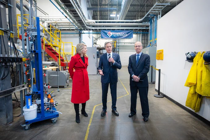 Secretary Blinken holds a microphone and speaks while standing in a warehouse at Lithion in Montreal. Foreign Minister Melanie Joly looks toward him as he speaks and a gentleman clasps his hands as he looks outward to the crowd. Equipment and protective gear line the walls, and a red staircase with yellow rails sits off in the background. A sign that reads "Lithion" hangs from pipes in the ceiling behind them.