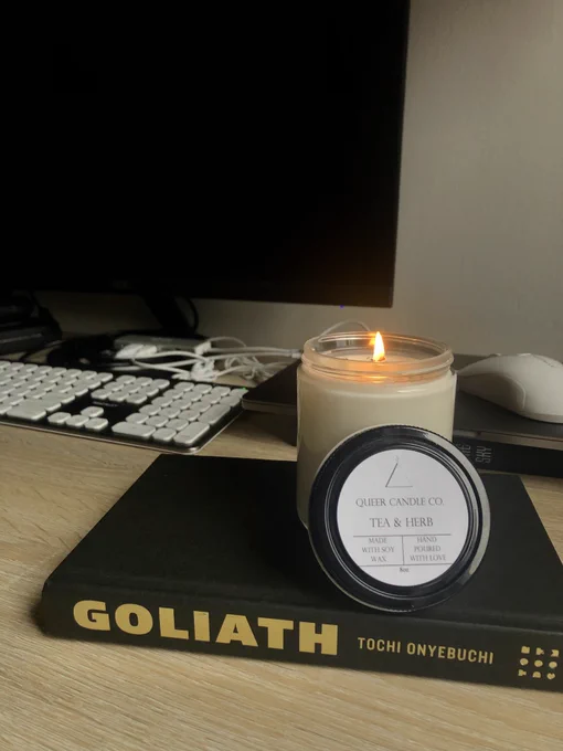 A hardcover copy of Goliath by Tochi Onyebuchi without its dust jacket that has a lit candle sitting on top of it. The candle is scented like Tea & Herb.