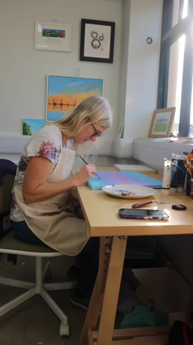 Sarah is sitting at her studio desk, leaning over a blue, lilac and pink painting of a sky. There is light shining in from a large window on the right.