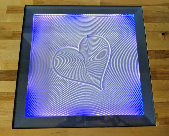 Wooden countertop with square hole replaced with glass. Under the glass is a blue lit pattern drawn in the sand by a steel marble. Robotic precision. The pattern is a heart that gets gradually larger while it twists slightly.