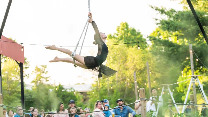 A performer from Parallel Exit hangs from an aerial setup above a gazing crowd. 