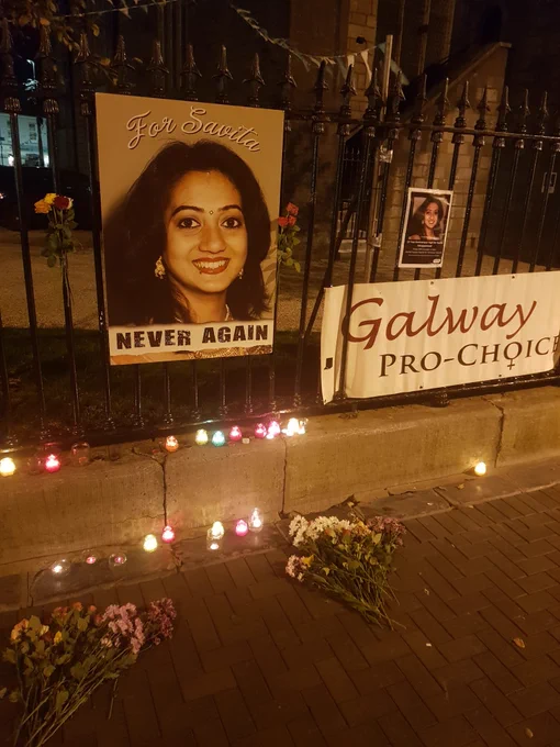 A tribute to Savita Halappanavar in Market Square, Galway. There are pictures of her and a Galway Pro-Choice banner on the railings next to the church. There are flowers and candles on the railings and on the ground.