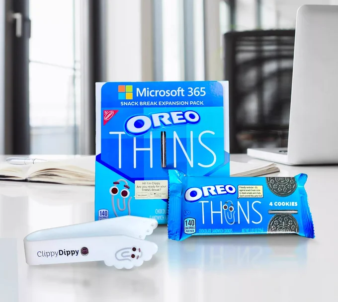 Blue packages of OREO THINS with an image of Clippy, a paper clip with eyes, sit on a desk next to a laptop and notebook.