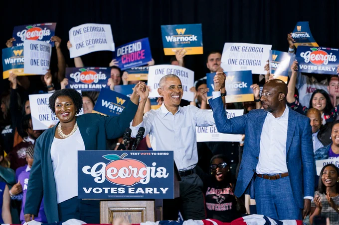 A photo of Stacey Abrams, President Barack Obama, and Senator Reverend Raphael Warnock holding hands and raising high at a rally in Atlanta, GA. A crowd of supporters standing behind them are also holding signs and cheering.