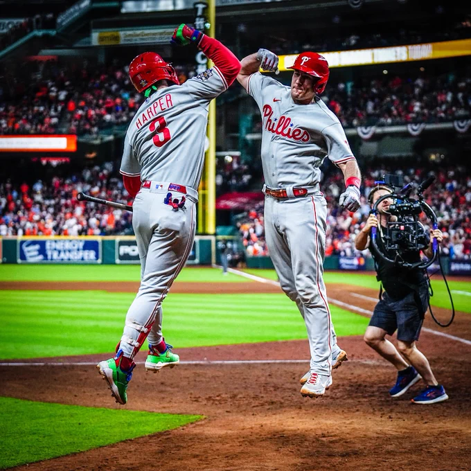J.T. Realmuto and Bryce Harper jumping in the air and celebrating Realmuto's game-winning home run in Game 1 of the World Series because they both know he's the best catcher in baseball. They're wearing the grey Phillies uniform.