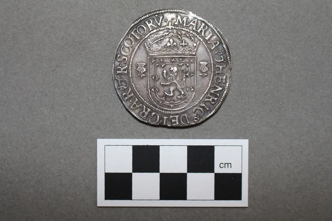 A silver coin from Mary Queen of Scots collection. It sits above a checkerboard ruler. 