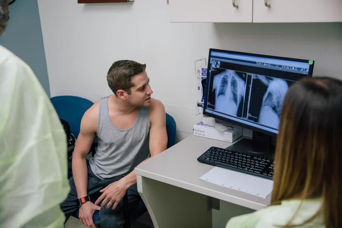 A light-skinned man sits and looks at a chest scan on a monitor with two medical professionals.