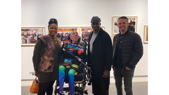 IMAGE DESCRIPTION 2: In front of some of Holly-Marie’s portraits of her debut solo exhibition hanging on a white wall at the Leica Store Mayfair, a landscape photo of Sulaiman next to his friends Holly-Marie, Simon, and Will. All are smiling and are joyful.