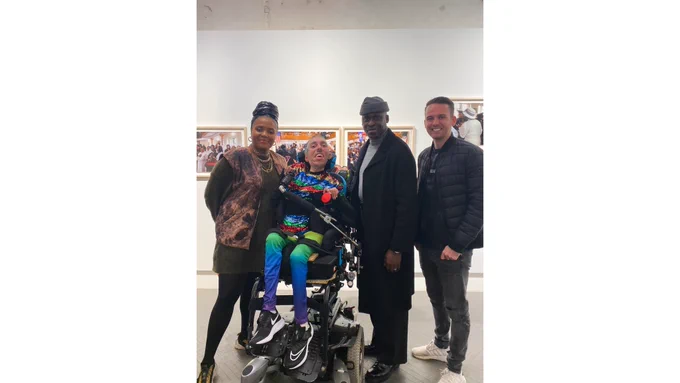 ID 1: In front of some of Holly-Marie’s portraits of her debut solo exhibition hanging on a white wall at the Leica Store Mayfair, a portrait photo of Sulaiman (a wholehearted Disabled AF & British-Pakistani man) sitting in his power wheelchair at full elevated height next to his friends Holly-Marie (a talented Black woman with gold hoop earrings and gold necklaces), Simon (a super creative Black man), & Will (a badass white man). Sulaiman is wearing his magnificent rainbow-coloured sequinned t-shirt with a black long-sleeved top underneath, his new Multicolour Daydream Meggings via @KapowMeggings, & his new Nike trainers. Holly-Marie is wearing a black leather head-wrap, a dark green long-sleeved top with a brown abstract print waistcoat, black trousers, & yellow trainers. Simon wears a grey beanie hat, a grey polo neck sweater, a black overcoat, & black shoes. Will is wearing a dark grey t-shirt with a black puffer jacket, grey jeans, & white trainers. All are smiling and are joyful.