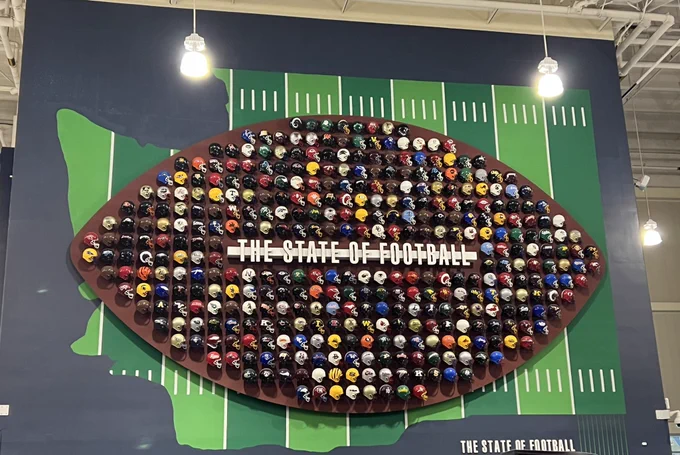 Large wall display with Washington State high school football helmets arranged in columns and rows in the shape of a football overlaying a map of the state with yardage markings.