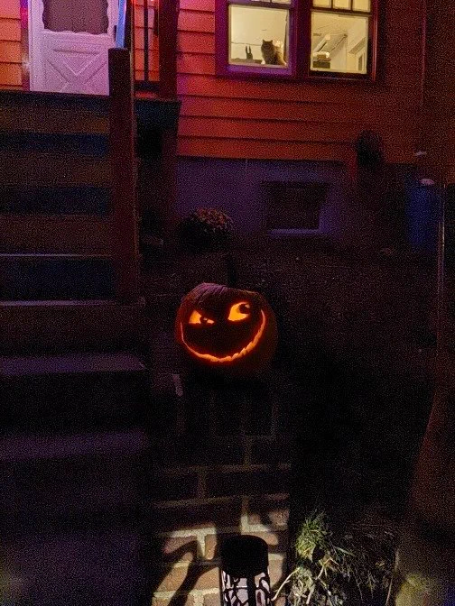 Leering pumpkin, carved into a jack-o'-lantern, one eye bigger than the other, lit from inside, sitting at the bottom of stairs. There is a cat watching from the window.