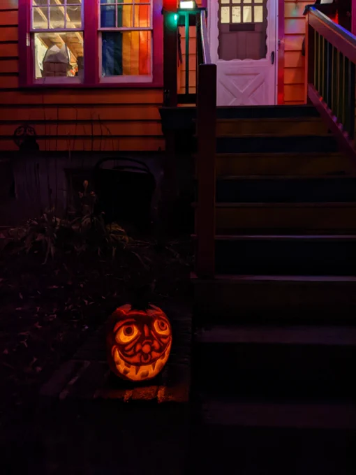 Pumpkin carved in relief into a grinning toothy jack-o'-lantern sitting at the bottom of stairs. There is a pride flag in the window.