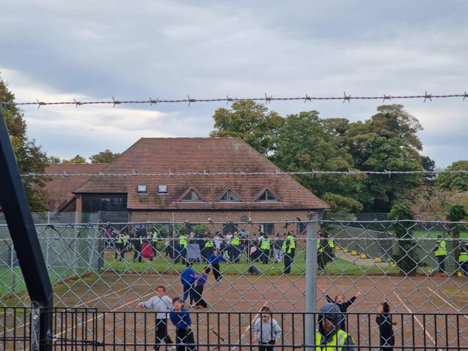 Children ar Manston camp behind barbed wire fences, with security and more detained people in tbe background