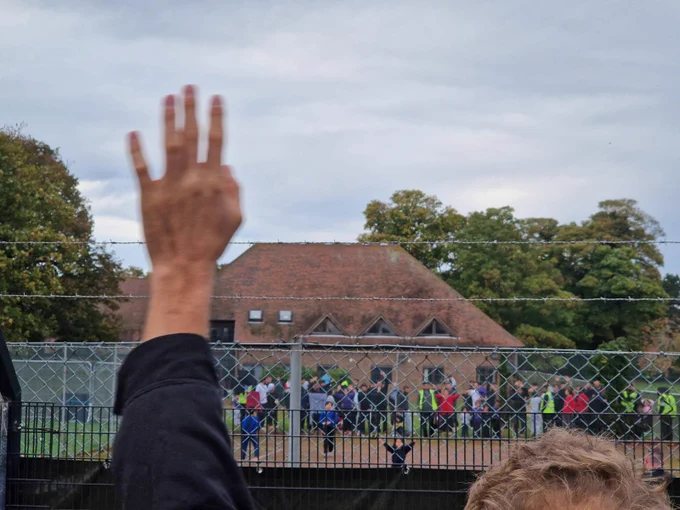 A protester waving to the people and children behind the barbed wire fence at Manston