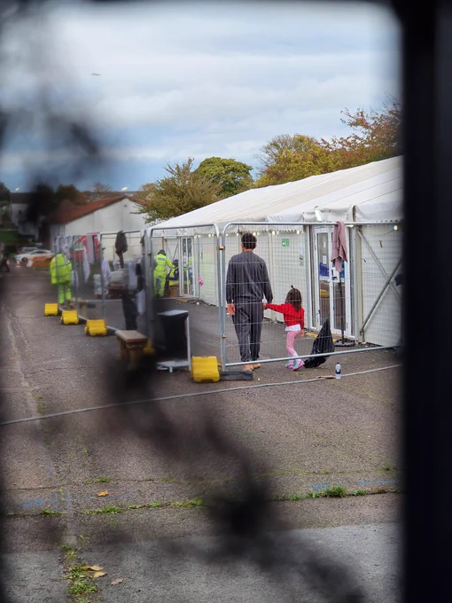 A man holding his child’s hand inside a fence cage at Manston camp. Security can be seen in the background.