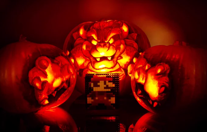 Pumpkin carving featuring Bowser from The Super Mario Movie. The design is carved across three pumpkins; one large one for Bowser’s face and body, and two smaller ones either side for his grabby hands. The design has been staged to look like Bowser is grabbing an 8-bit version of Mario.