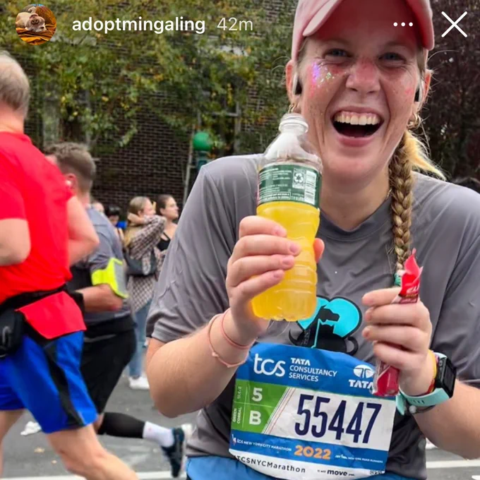 A young woman with reddish-blonde hair in a braid laughs at the camera while holding an orange drink and wearing a PAWS NY shirt with her TCS NYC Marathon bib: #55447. She is wearing a pink hat and has sparkles on her face. The image is a screenshot from Instagram, with the handle @adoptmingaling in the corner.